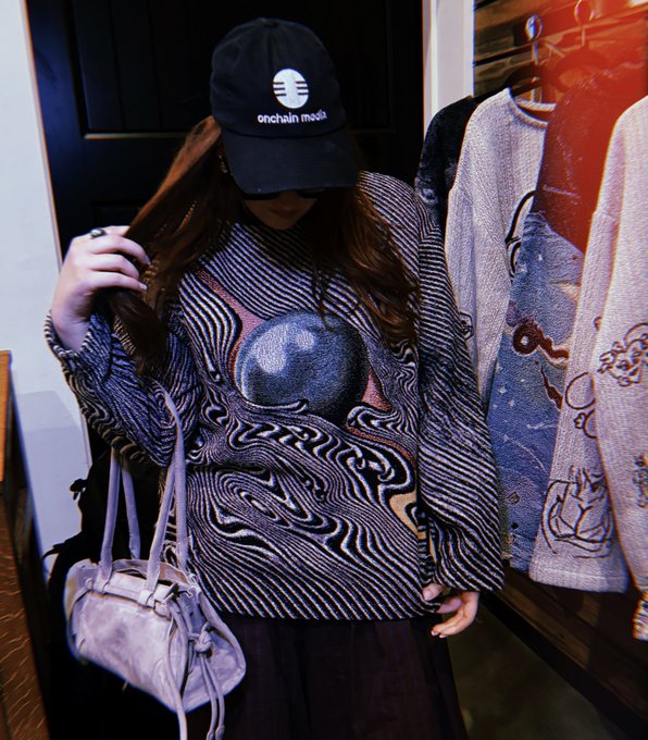 A person wearing a black 'onechain mode' cap and a patterned long-sleeve top with a large moon design holds a purple handbag.