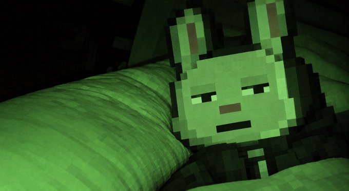 A pixelated, Minecraft-style green creature with pointed ears lies on a green pillow, viewed in dim lighting.