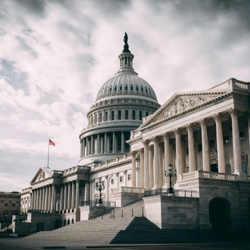 The U.S. Capitol Building stands under a cloudy sky, with the American flag flying prominently beside its iconic dome and columns. The image underscores the post's reference to the CLARITY Act's Senate procedural vote scheduled for 2:15 pm ET today, as Polymarket odds of the legislation being signed into law have dropped to 19%.