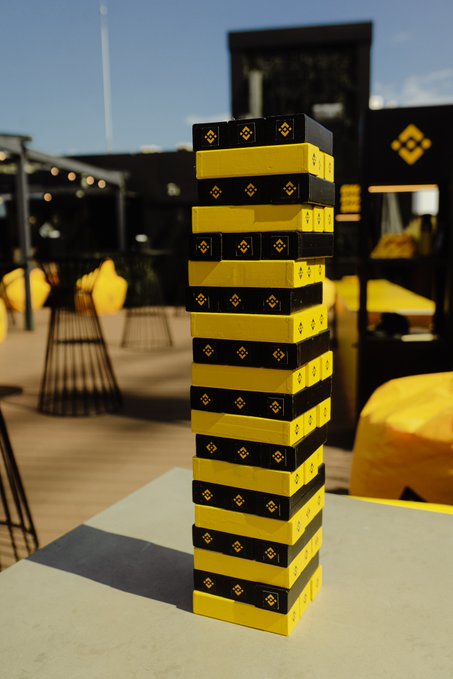 A tall, carefully balanced Jenga tower features black and yellow blocks prominently displaying the Binance logo, sitting on an outdoor table. The image ties into the post's caption by showing a leisurely Saturday afternoon game, likely at a Binance-branded event or lounge.