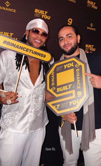 Two men pose smiling at Binance's 9th anniversary celebration in Dubai, holding branded props including a "#BINANCETURNS9" sign and a "Built By You" logo, with the "BUILT BY YOU" and "BINANCE" branding visible on the backdrop behind them.