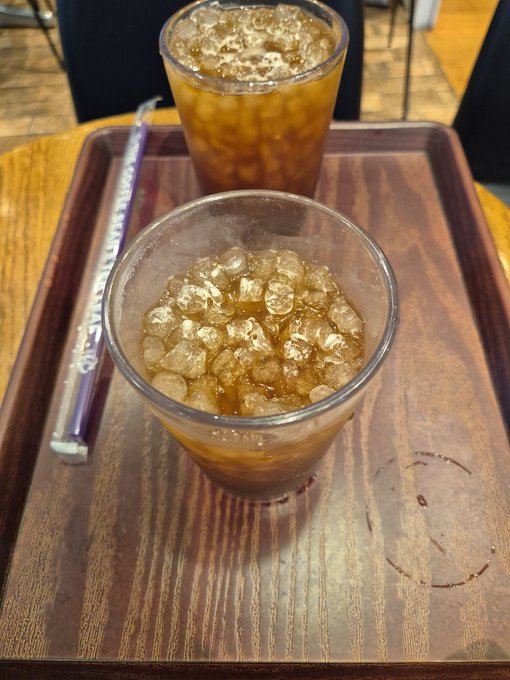 Two clear glasses of iced coffee or a similar brown beverage on a wooden serving tray, with a wrapped straw placed beside them.