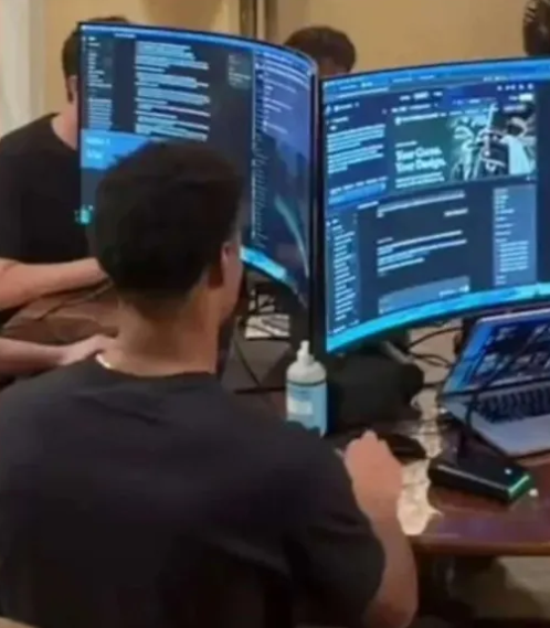 A person works at a desk with multiple curved monitors displaying lines of code, while another person sits nearby in a dimly lit room.