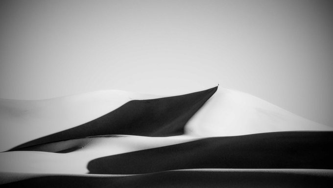 A minimalist black-and-white photograph of smooth sand dunes, with a single tiny figure standing at the crest of a ridge beneath a pale sky.