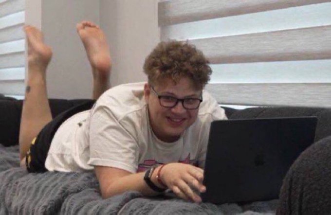 A smiling young man with curly hair and glasses lies on his stomach on a bed, using a laptop with his feet kicked up behind him.