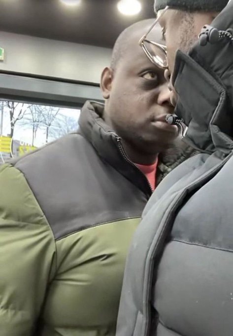 A close-up selfie of a man in a green and black puffer jacket on public transit, giving an intense side-eye glance to another passenger standing next to him.
