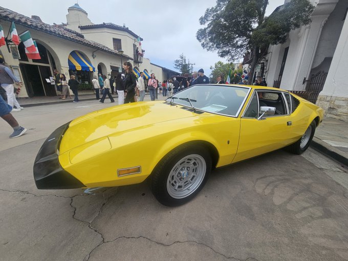 A bright yellow De Tomaso Pantera sports car parked on a street in front of a building with Spanish-style architecture and Italian flags, with pedestrians in the background.