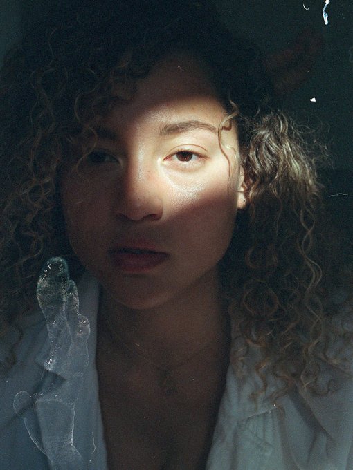 A close-up, moody portrait of a young woman with curly hair, wearing a light blue shirt with a translucent plastic-like object near her shoulder.