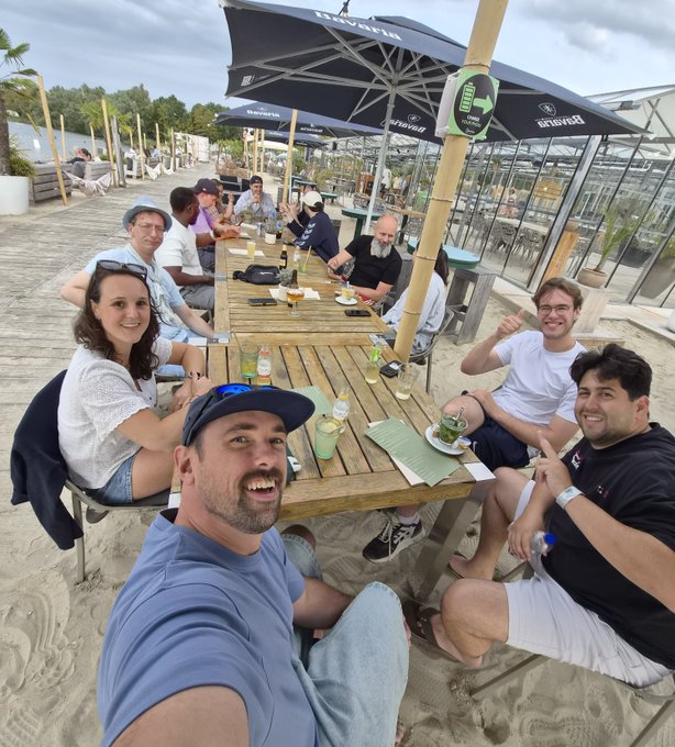 A group of people sitting at a long wooden table outdoors at a beach venue under umbrellas, posing for a photo.