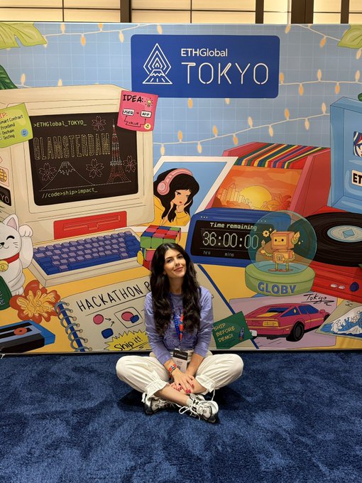 A young woman sits cross-legged on the floor in front of a vibrant "ETHGlobal TOKYO" mural featuring retro tech illustrations, with a digital timer reading "36:00:00" remaining and a lucky cat figure nearby, reflecting day 2 of the hackathon experience.