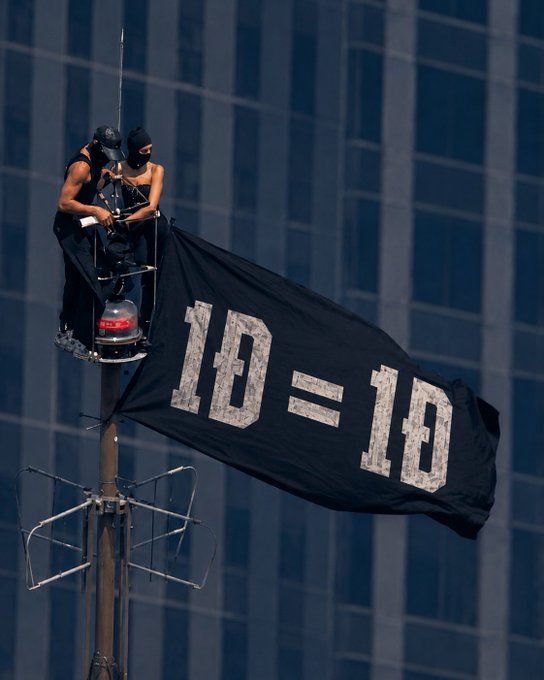 Two masked individuals perch on a tall pole displaying a large black flag reading "1D = 1D" against the backdrop of a glass office building.