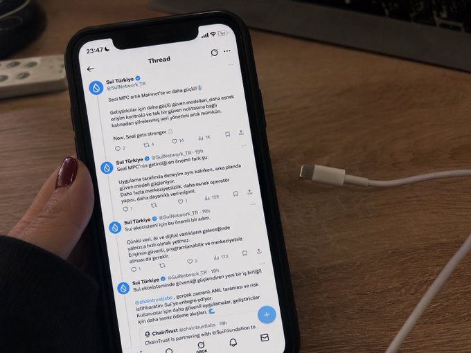 A hand with dark nail polish holds a smartphone displaying a "Thread" on X (formerly Twitter) with multiple posts from "Sui Türkiye" (@SuiNetwork_TR), while a white charging cable lies unplugged nearby on a wooden desk. The caption aligns with the post's theme, as the phone shows a battery level of just 1% next to the active Sui network thread.