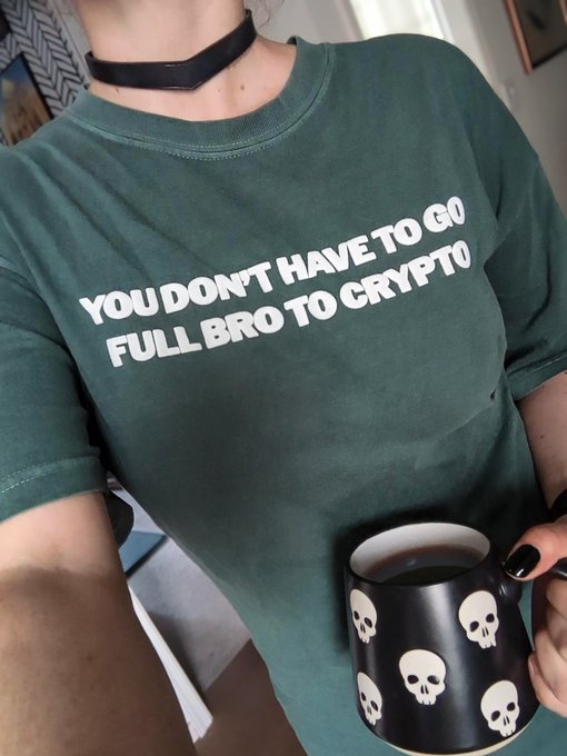 A person wears a dark green t-shirt with white text reading 'YOU DON'T HAVE TO GO FULL BRO TO CRYPTO', holding a black mug decorated with white skulls.
