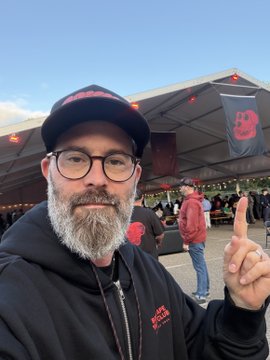 A selfie of a bearded man with glasses wearing a black hoodie and cap, pointing upward at a 'M3' banner in the background.