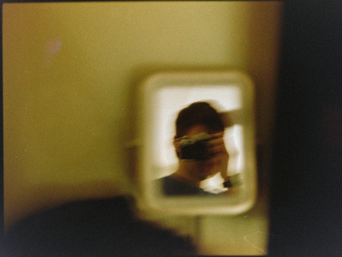 A blurry, vintage-style photograph showing a person taking a mirror selfie with a camera, reflected in a square mirror mounted on a wall.