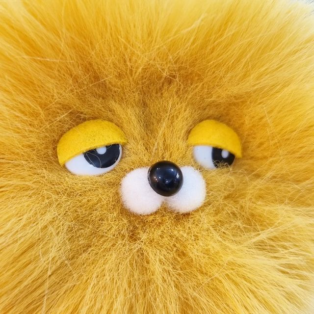 A close-up of a fluffy yellow stuffed animal's face featuring large cartoonish eyes with yellow eyelids and a black nose.