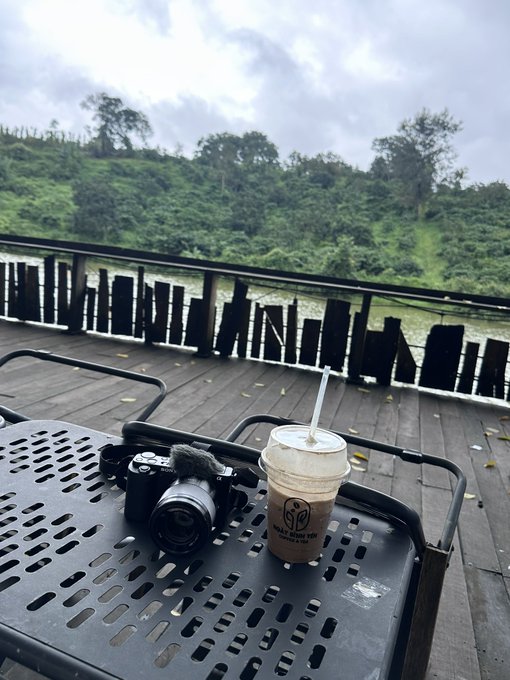 A Sony camera and an iced coffee sit on a black metal table overlooking a wooden deck and lush green hillside. The setup pairs with the post's nostalgic reflection on a missed Bitcoin opportunity, evoking a moment of quiet contemplation over past regrets.