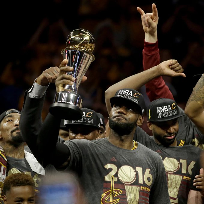 LeBron James holds the NBA Finals MVP trophy aloft while wearing a "2016 CHAMPS" shirt and cap, celebrating the Cleveland Cavaliers' championship victory. The image captures the historic moment referenced in the post about "The Greatest Basketball Player of All Time," with teammates around him also sporting championship gear.