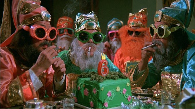 Several figures dressed as gnomes with white beards, ornate robes, and oversized goggles gathered around a green birthday cake with a lit '6' candle, with one smoking a cigarette.