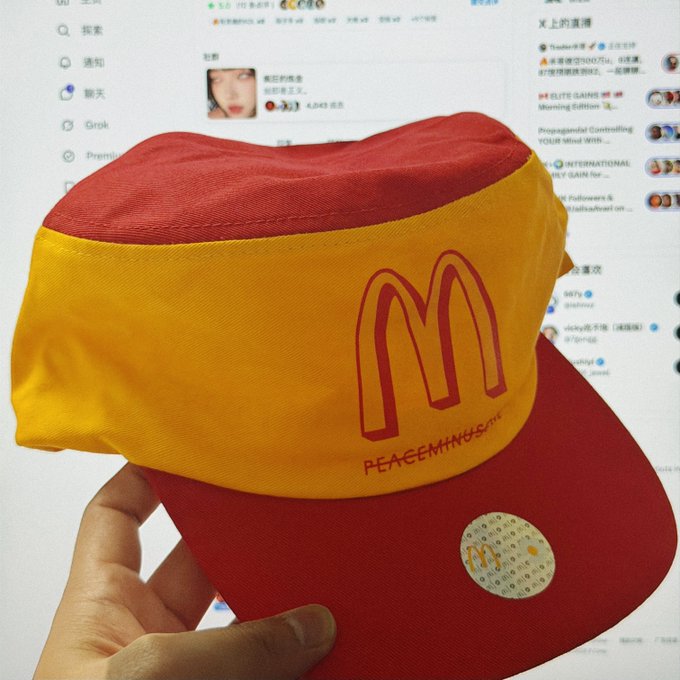 A hand holds up a red and yellow McDonald's 'PEACE MINUS ONE' branded visor hat in front of a computer screen displaying social media.