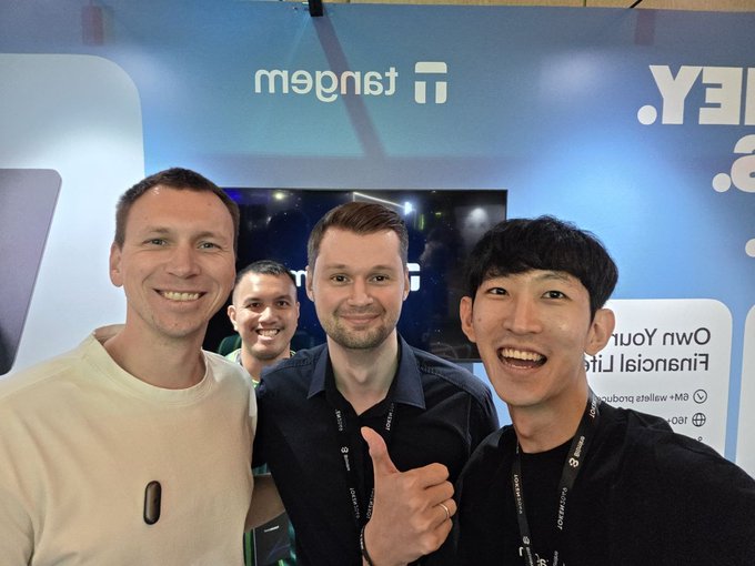 A selfie of four men at a crypto event booth, with Tangem branding and “Token2049” visible on the blue backdrop alongside event details such as “Owned Your Financial Life” and “PB4-48.”