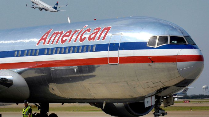 An American Airlines jet with "American" branding in red sits on the tarmac, while another aircraft flies in the background, with a ground crew member visible near the landing gear. The image relates to the "FAA = HBAR" post by symbolizing U.S. aviation regulation, representing the aircraft and airport infrastructure overseen by the Federal Aviation Administration.