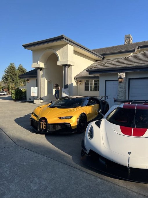 Two Bugatti hypercars—one yellow with the number 16 and one white with red stripes—parked in the driveway of a Mediterranean-style mansion.