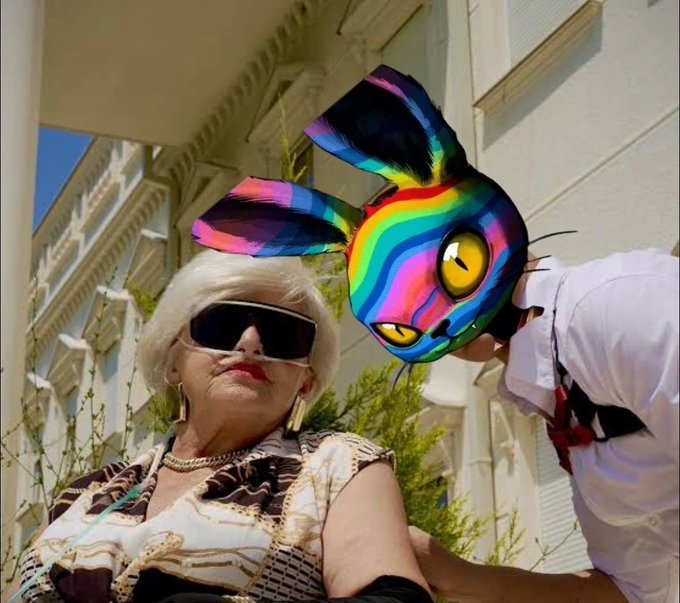 A stylish woman with white hair and dark sunglasses poses alongside a companion wearing a vibrant, rainbow-striped bunny mask in front of a sunlit building. The colorful, whimsical mask complements the cheerful "GM (good morning)" greeting, adding a playful "Plan D" twist to the upbeat message.