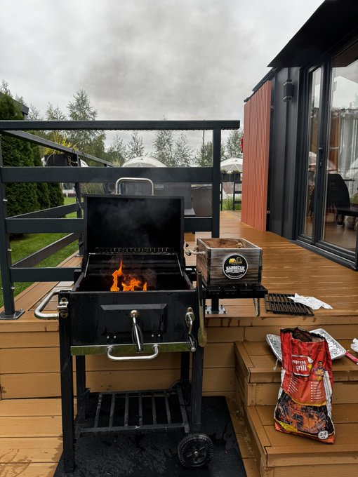 A black charcoal grill with flames is set up on a wooden deck next to a bag of "Cowboy" brand charcoal (24 lbs), with smoke rising against a moody overcast sky — perfectly capturing the laid-back backyard cookout "+ vibe."