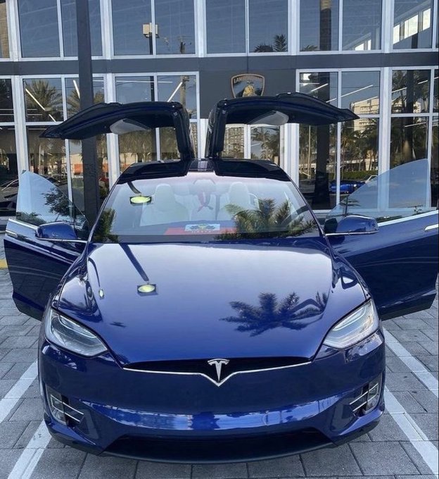 A dark blue Tesla Model X is parked in front of a Lamborghini dealership with its falcon-wing rear doors and front doors open, displaying the iconic Tesla logo on the front.