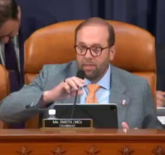 The image shows a man sitting at a wooden desk with a microphone in front of him while he speaks into it, possibly addressing a House Committee meeting or participating in a hearing related to Bitcoin tax reform. The scene suggests that the man might be discussing the impact of Bitcoin on the U.S. economy and how it affects traditional financial systems.