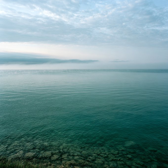 A serene seascape featuring calm, clear turquoise water in the foreground and a misty, cloud-covered horizon under a pale sky.