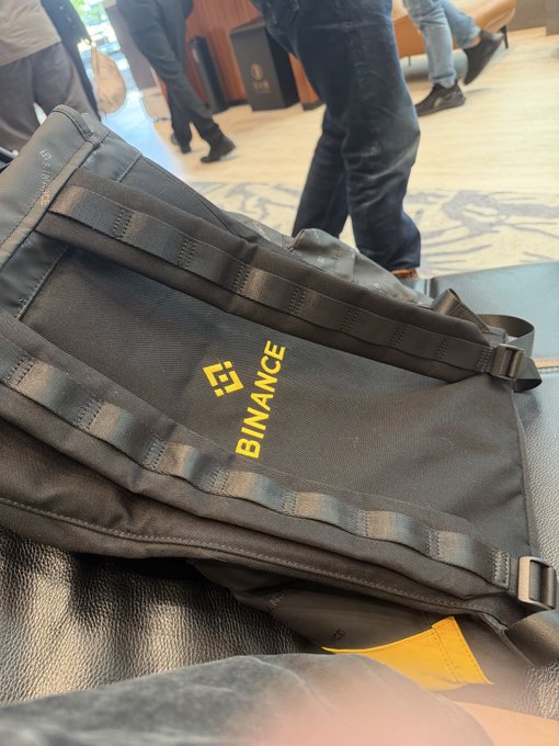 A dark gray tactical-style backpack with yellow 'BINANCE' branding sitting on a black leather chair.