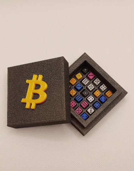 A dark gray foam display box features a raised yellow Bitcoin "₿" logo on the lid, sitting beside the open base which contains a grid of colorful dice in various colors.