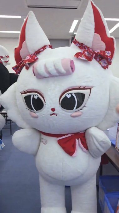 A white fox mascot costume with large expressive eyes and red floral bows on both ears stands in what appears to be a workshop space, matching the "JEWEL #GMMTVPOPUPSTOREINFUKUOKA2026" reference to the adorable character with side bows.