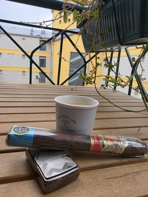 A still-life scene on a wooden balcony table features a cup from 'BRASSERIE LIPP ST-GERMAIN', a packaged cigar, and a black ashtray, with a view of yellow and white buildings through a railing.
