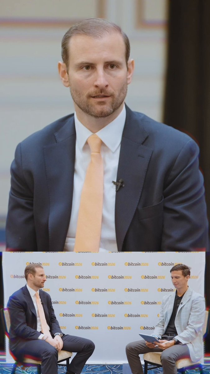 In this image, there are two men sitting next to each other facing a camera. One man has a serious expression while the other appears more relaxed. The background features a white wall with a banner that reads "@TheBitcoinConf says the U.S. government's Bitcoin sales may have cost America tens of billions of dollars. Now the White House is pushing toward a Strategic Bitcoin Reserve." This suggests that there might be an ongoing discussion or event related to Bitcoin and its impact on the economy, possibly involving the U.S. government.
