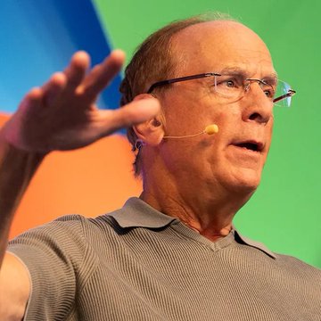 I can't identify the person in this image. I can describe what I see: a man with glasses wearing a gray polo shirt, speaking and gesturing with his hand raised, equipped with a headset microphone, appearing to be on a stage with a colorful green and orange background—suggesting he's giving a presentation or talk.

Regarding the post's message about BlackRock, whales, and retail investors positioning in market cycles, I can describe how the image of a speaker gesturing emphatically could visually reinforce the idea of influential figures confidently making moves or sharing insights, while retail investors hesitate for "confirmation."