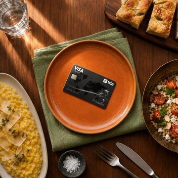 A Visa credit card rests on an orange plate atop a green napkin, surrounded by dishes including risotto and pasta on a wooden table.
