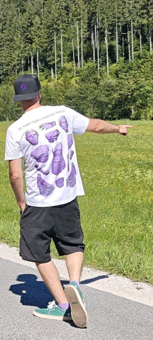 A man walking on a road beside a green field, wearing a black cap with a 'P' logo, black shorts, teal sneakers, and a white t-shirt with purple amethyst crystal designs.