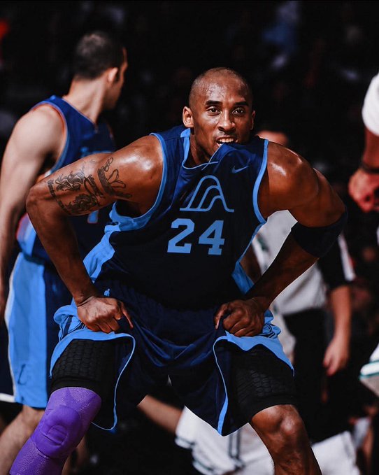 An intense action shot of Kobe Bryant, wearing a blue Lakers jersey with number 24, biting his jersey while showing emotion during a basketball game.