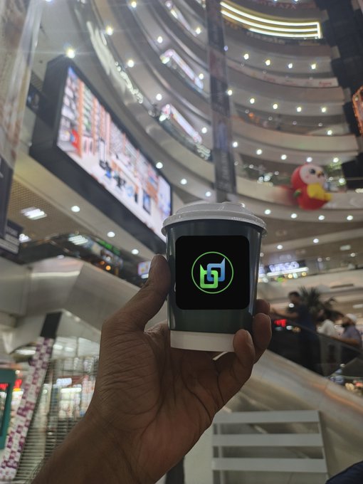 A hand holds a paper cup with a green 'T2T' or 'LST' logo in the foreground of a multi-story shopping mall interior.