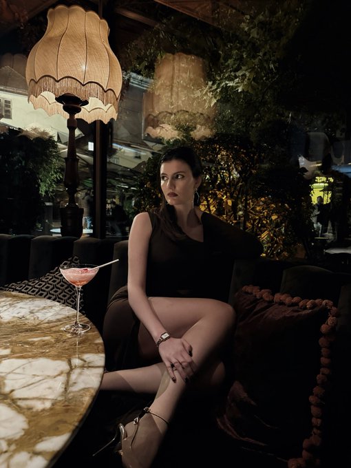 A young woman in a black dress sitting on a dark velvet sofa next to a marble table with a cocktail glass, lit by a vintage fringed lamp in a moody bar or lounge setting.