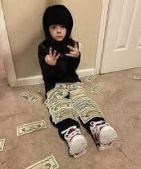 A young child wearing a black hoodie and pants sits on a carpeted floor, covered in dollar bills with more bills scattered around them, posing with hand gestures.
