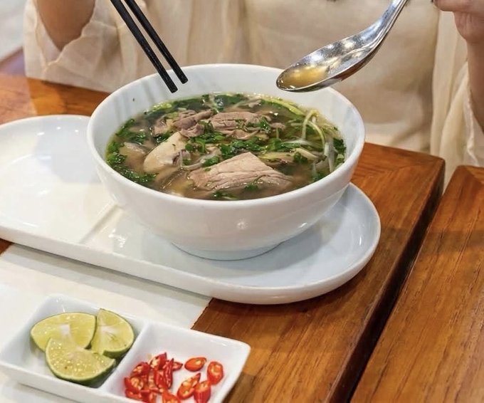 A bowl of Vietnamese beef noodle soup (pho) topped with herbs and bean sprouts, served with a side of lime wedges and sliced red chilies on a wooden tray.