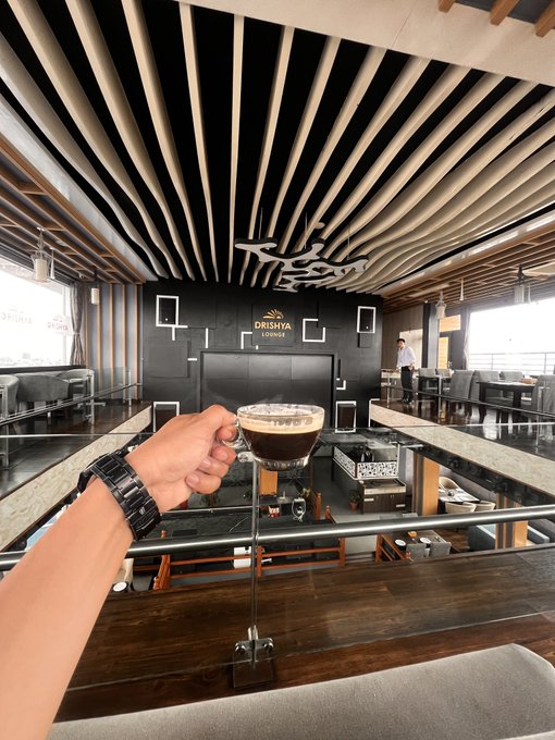 A hand wearing a black watch holds up a glass of espresso in a modern lounge area featuring wooden slatted ceilings and a 'DESSAVA LOUNGE' sign in the background.