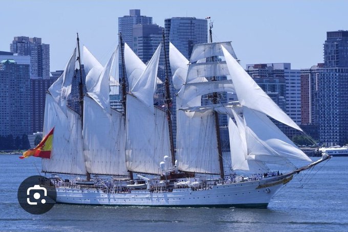 A large white tall ship with multiple masts and sails unfurled sails past a city skyline, flying a Spanish flag.