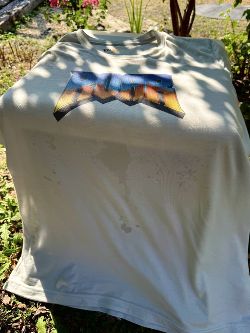 A white t-shirt with a metallic orange and blue logo lies in a garden setting, featuring a water stain across the front.