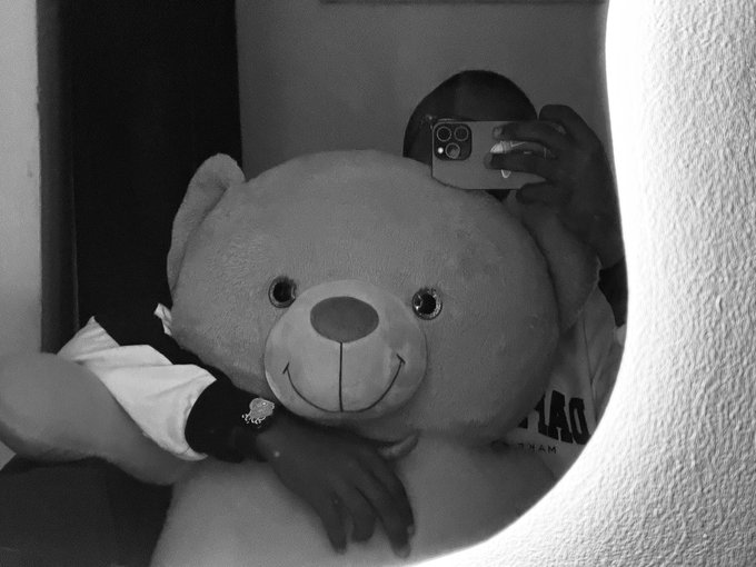 A person takes a mirror selfie while hugging a large teddy bear, using a smartphone to capture the moment in black and white, humorously suggesting that the stuffed companion is the highlight of their day—matching the caption "i love my job."