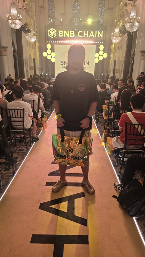 A man stands on a runway at a BNB CHAIN Summit event, holding a shiny gold and green BNB CHAIN tote bag, with the runway marked with letters spelling out part of "BNB."
